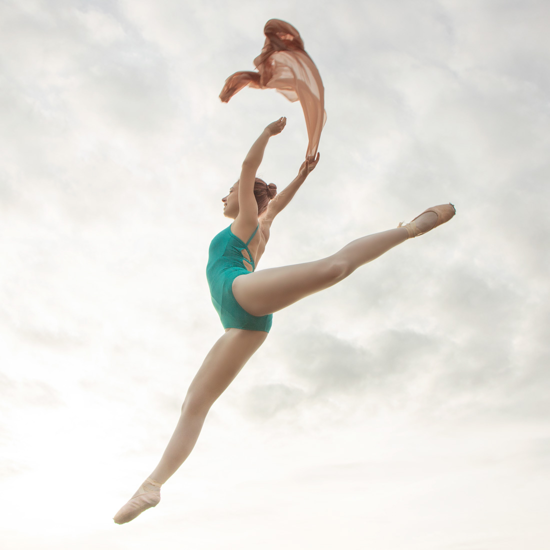 Ballerina in a turquoise leotard is jumping while holding a beige colour scarf. Creative ballet dance photography