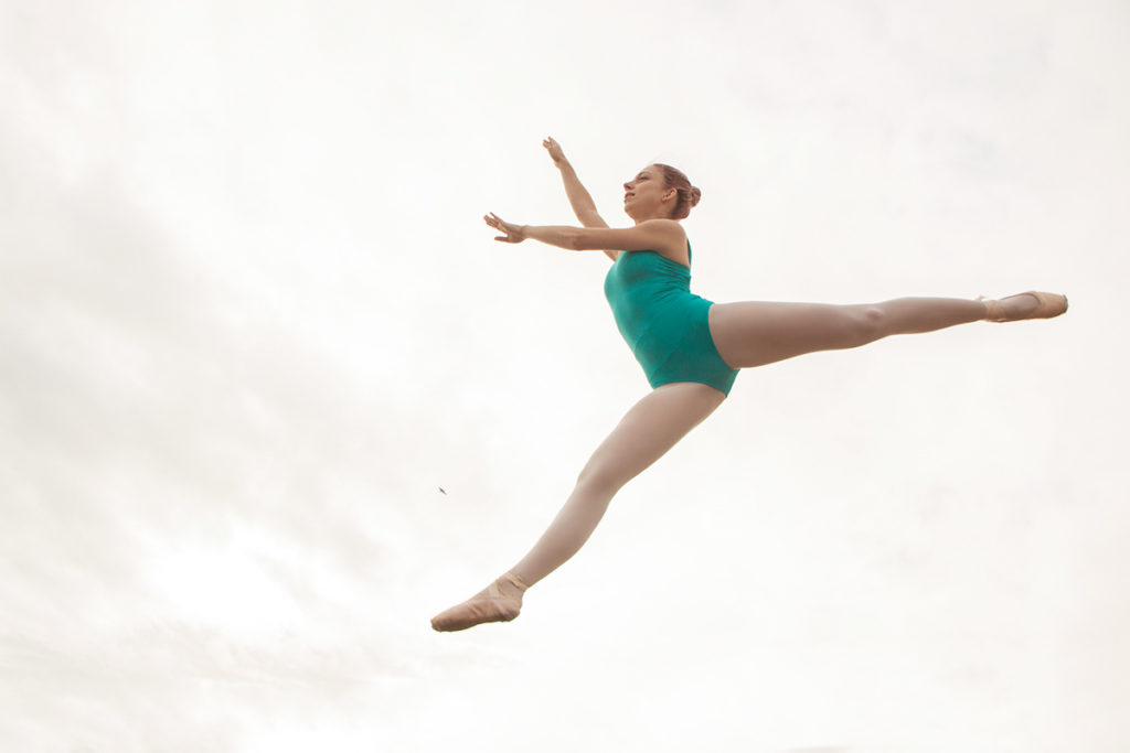 A ballet dancer wearing turquoise leotard is striking a pose in the air