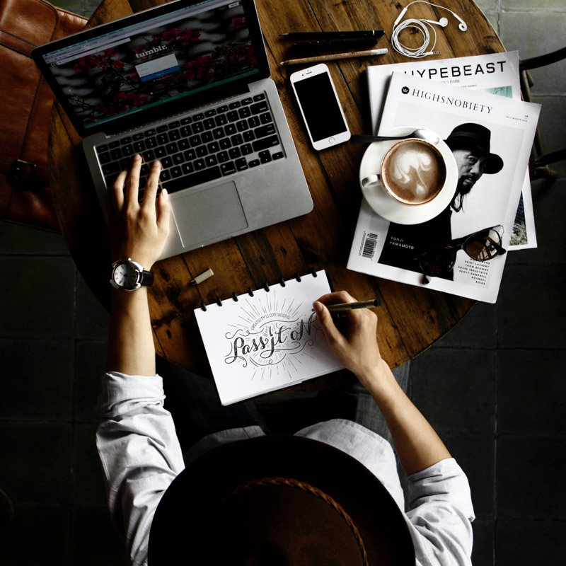 being productive when working from home. A man is drawing and searching internet. Cappuccino on a pile of magazines, phone and earphones on the table 