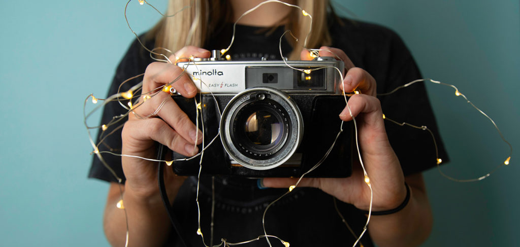 A blond woman is standing in front of blue wall, holding Minolta film camera ready to take a photo, with fairy lights surrounding camera.