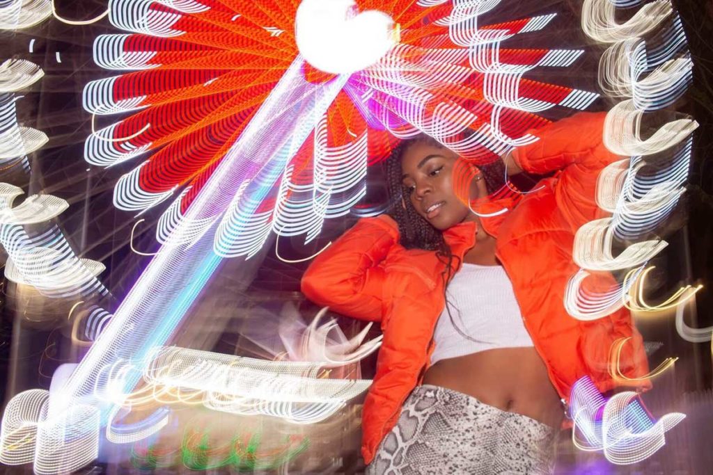 A black woman is dancing in front of Ferris wheel at night. she is wearing red jacket, white top and snake leggings. Slow sync flash creates interesting effect of light painting 