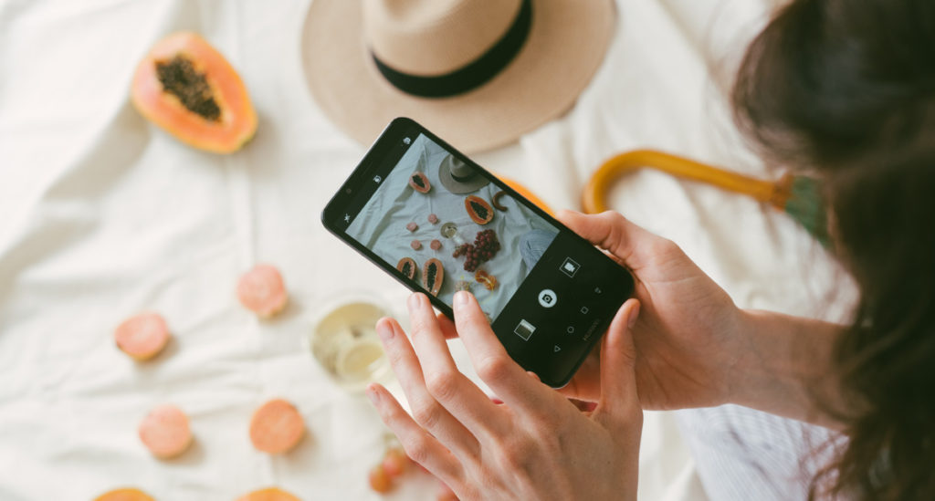 A woman is taking photo of fruit and beige hat on white bed sheet. 