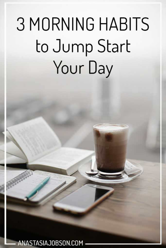 A wooden table with an open book, planner with a blue pen lying on top, a glass of cappuccino and a phone. Text saying 3 morning habits to jump start your day. 