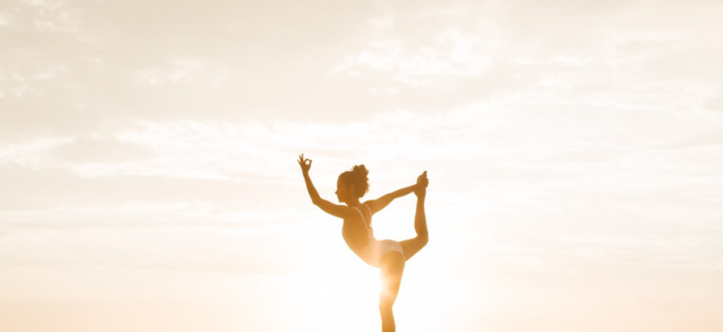 Woman wearing a swimsuit is striking yoga dancer's pose during golden hour with sky as a background