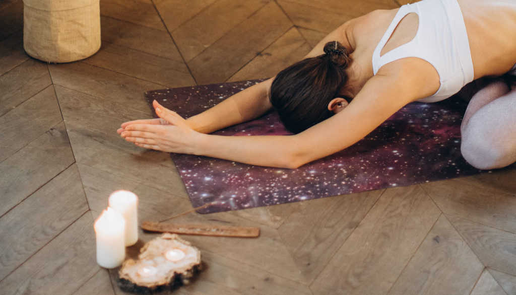 A woman wearing a white sports top is practicing yoga on a wooden floor