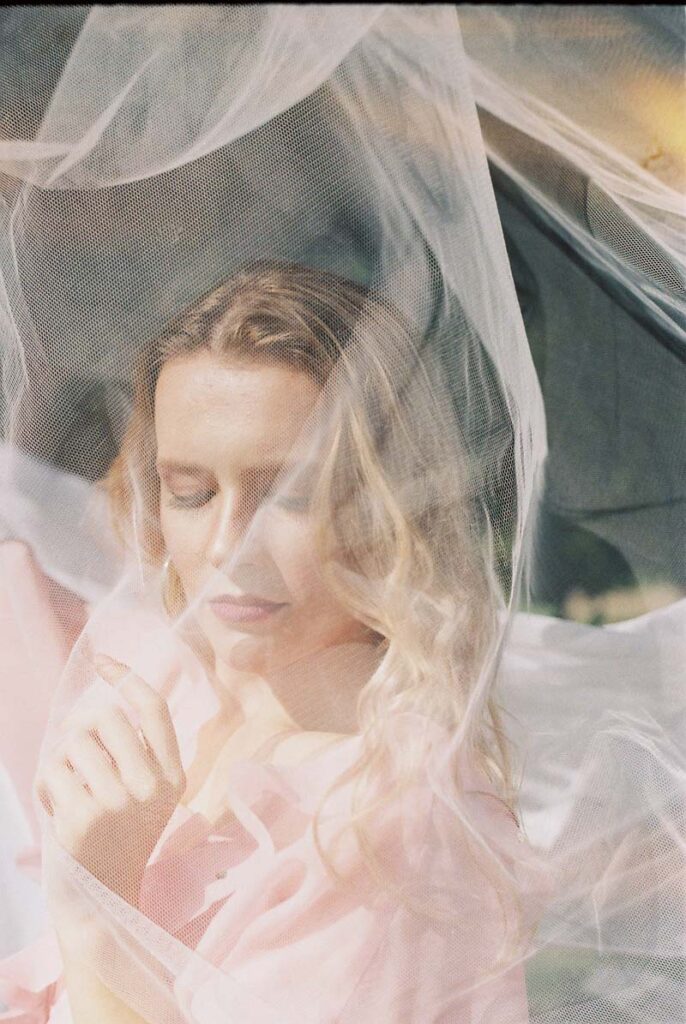 35mm portrait, film photo inspiration. A portrait of a young woman in pink dress and wavy hair behind a white net. Outdoor portrait photoshoot in Birmingham U.K.