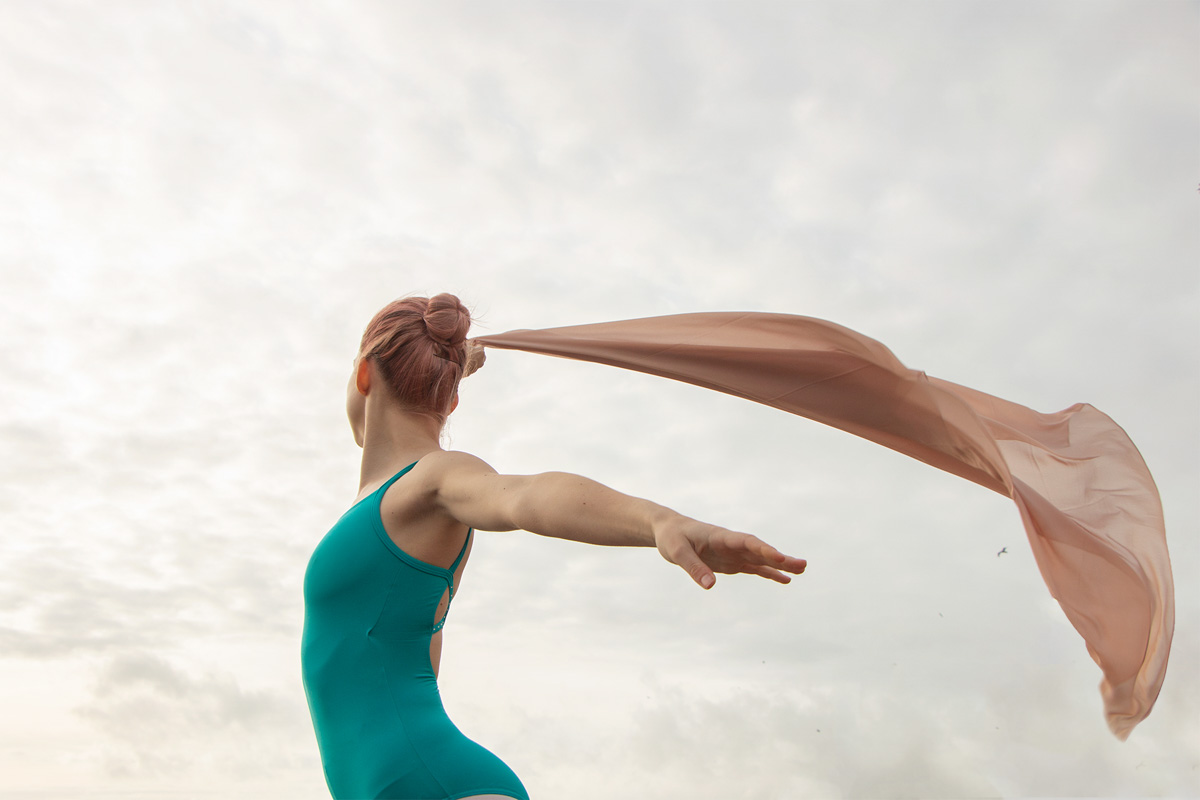 Female ballet dancer wearing a turquoise bodysuit is dancing with beige fabric. Ballet dance photoshoot on the beach. Creative dance photography